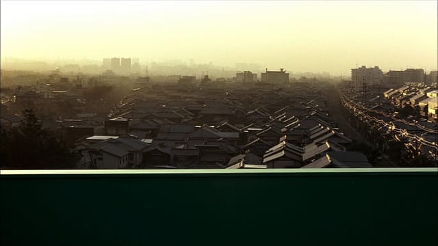 夜明けの高い位置から見下ろした、霞んだ低層の街並み。手前の暗い緑の手すりの向こうで、近くの屋根に朝日が当たりはじめている
