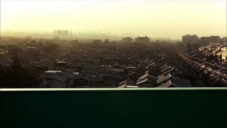 夜明けの高い位置から見下ろした、霞んだ低層の街並み。手前の暗い緑の手すりの向こうで、近くの屋根に朝日が当たりはじめている