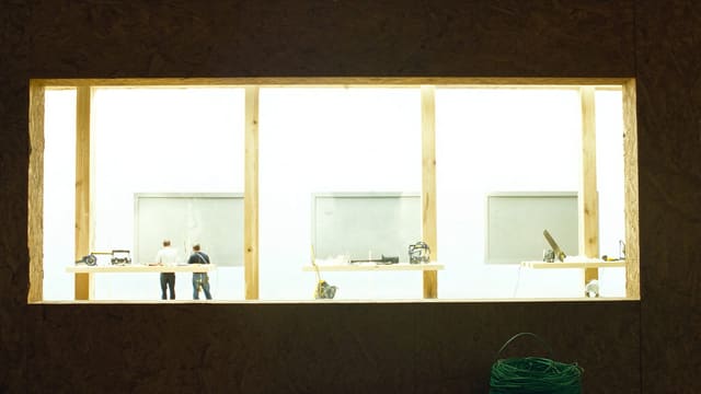 内装工事中の現場。大きな開口から光が差し込み、奥に二人の作業者が小さく見える。手前に緑のケーブルが巻かれている