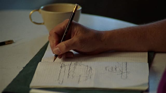 方眼ノートに鉛筆で細かな印を書き込んでいる手元。奥に生成りのカップが置かれている