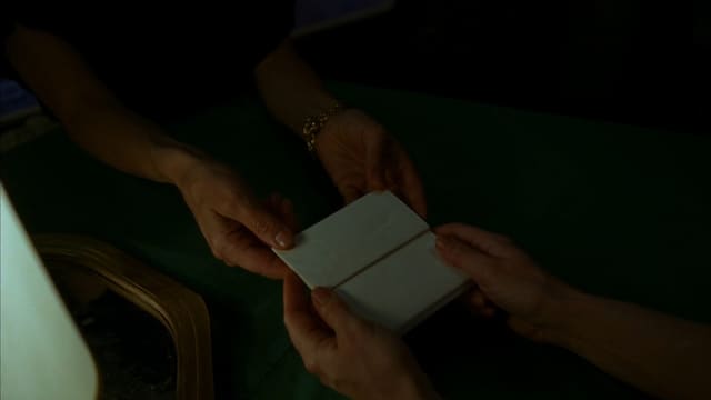 濃い緑の卓上で、二組の手が小さな白い折りカードを受け渡している手元の接写