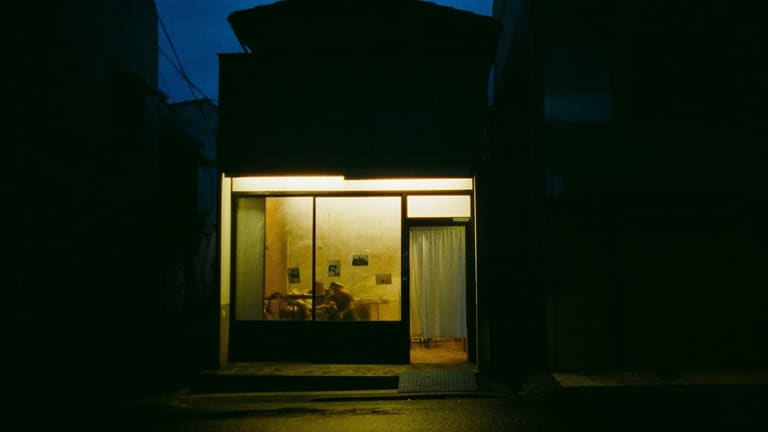 夕暮れの通りに面した小さな店。中の明かりだけが灯り、暖かい光が路面にこぼれている