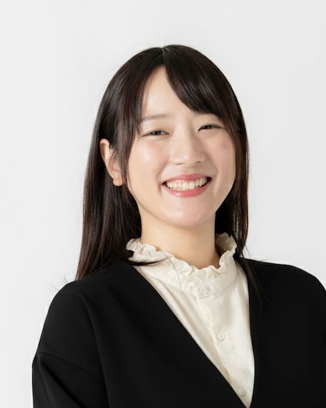 白い背景の前で、黒いジャケットを着てこちらを見ている西本優子