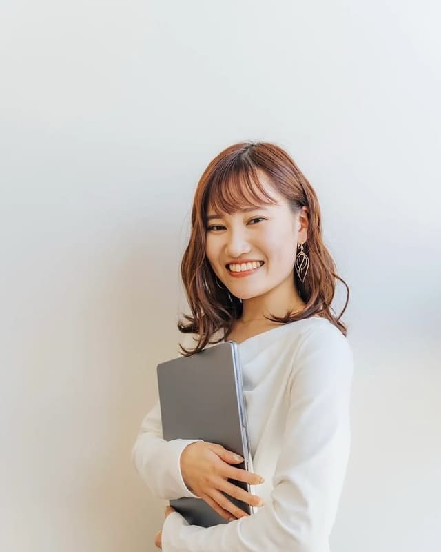 白い背景の前で、ノートパソコンを抱えてこちらを見ている坂本あすか